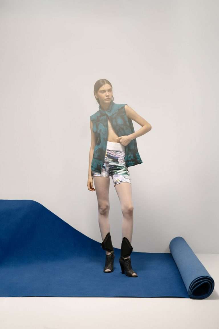 Una mujer, de pie sobre una colchoneta azul con el borde enrollado, lleva una chaqueta estampada sin mangas, pantalones cortos de ciclista estampados y botas negras con puntera abierta. El fondo es liso y claro, creando un ambiente de estudio minimalista.