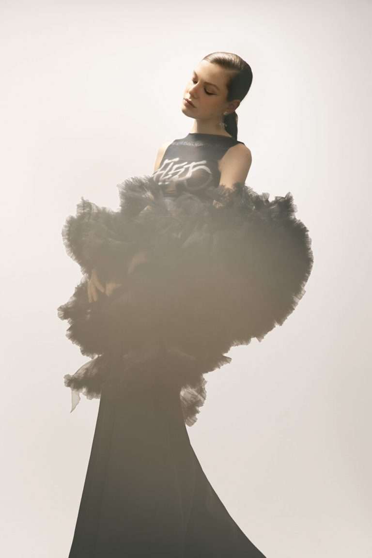 Una mujer con un espectacular vestido negro de voluminosos volantes de tul, de pie sobre un fondo claro, mira hacia abajo con el pelo recogido en una coleta baja. La imagen tiene una iluminación suave y etérea.