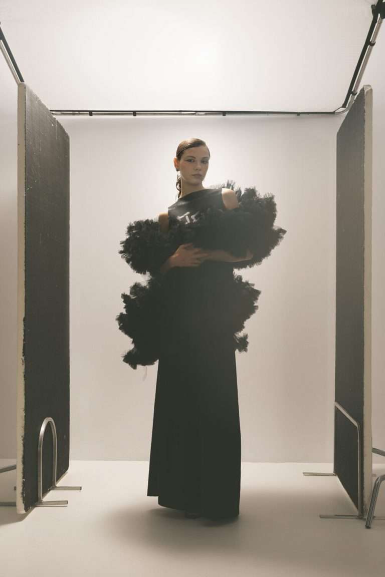 Una mujer con un elegante vestido negro con espectaculares detalles de tul negro se sitúa con confianza entre dos grandes paneles verticales en un estudio bien iluminado.