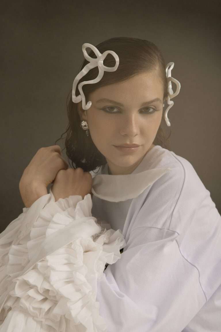 Una mujer con el pelo castaño mojado lleva esculturales accesorios blancos para el pelo, un top blanco con volantes y pendientes de perlas, y posa con los brazos cruzados sobre un fondo oscuro.