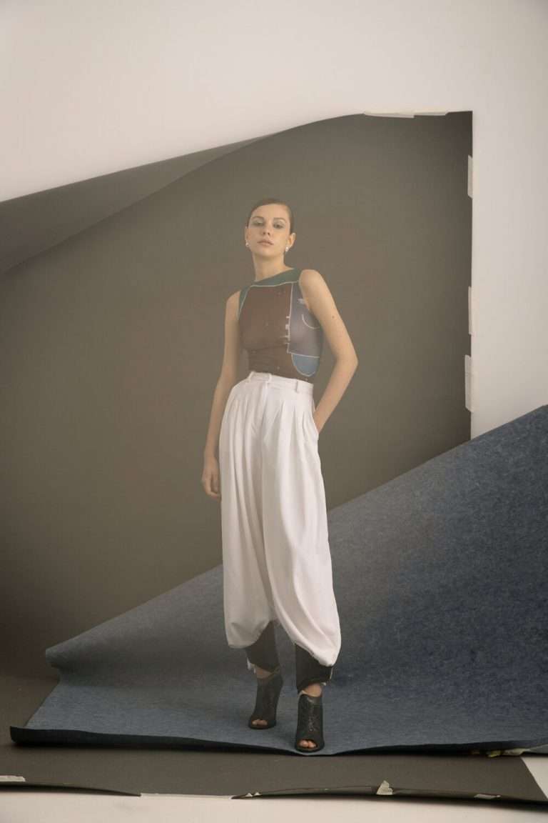 Una mujer de pie delante de un fondo de estudio abstracto, con un top estampado sin mangas, pantalones blancos sueltos de cintura alta y zapatos negros abiertos, con una mano en el bolsillo y una expresión neutra.