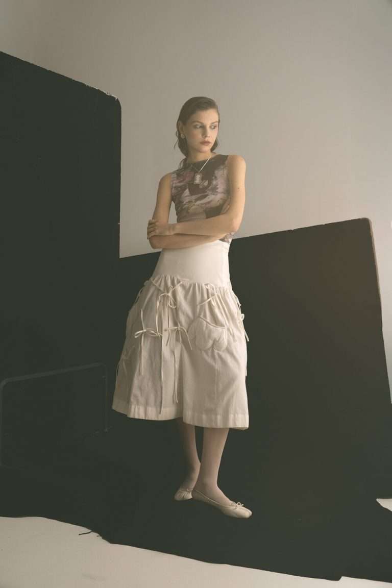 Una mujer de pie con los brazos cruzados, vestida con un top estampado sin mangas, una falda midi blanca con detalles de volantes y unos zapatos planos de color crema, posa delante de unos fondos de estudio minimalistas en blanco y negro.