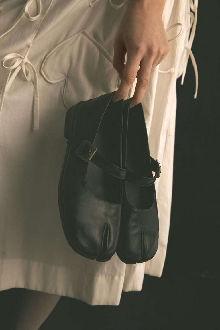 Una persona vestida de blanco sujeta por las correas unos zapatos Tabi de cuero negro con diseño de tiras y puntera partida, sobre un fondo oscuro.