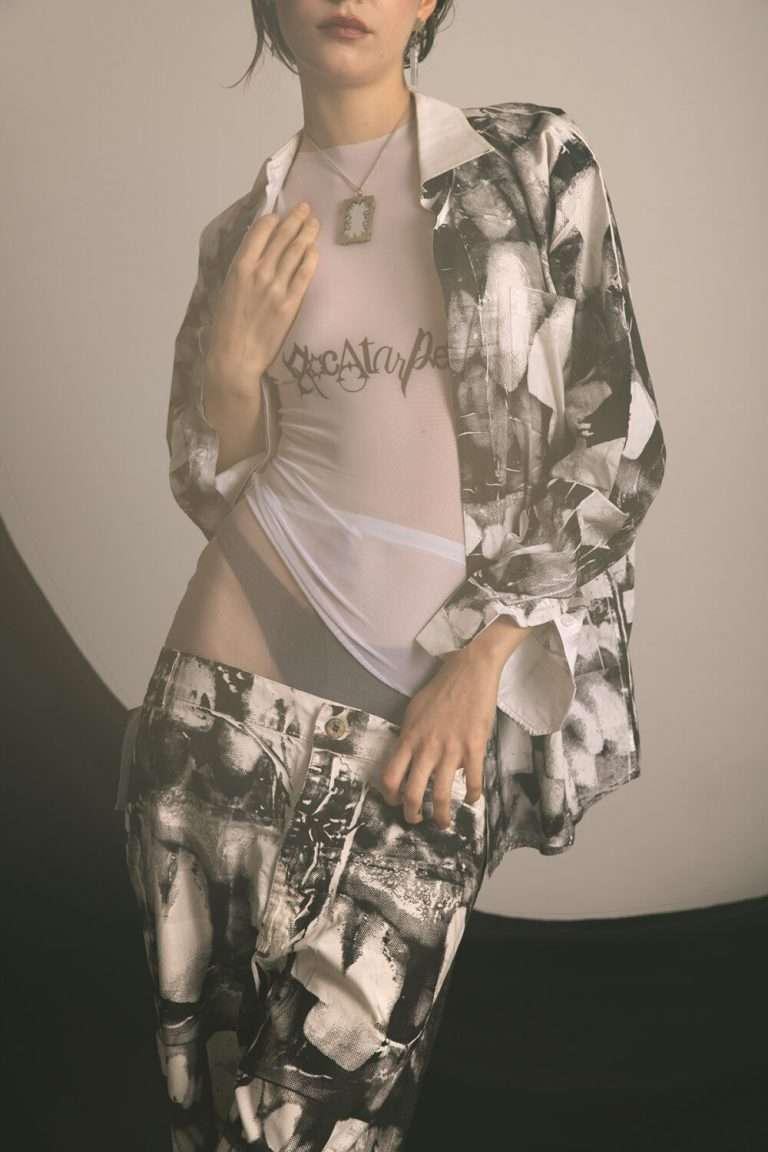 Persona vestida con pantalones sueltos estampados en blanco y negro y una camisa abierta a juego sobre un top blanco transparente con texto. Lleva el pelo corto y oscuro y un collar camafeo. El fondo está suavemente iluminado y es neutro.