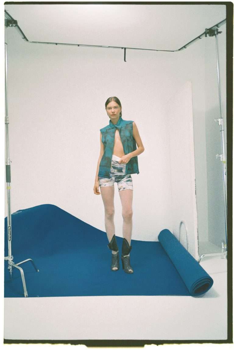 Una persona de pelo castaño liso está de pie sobre un fondo azul en un estudio, con un chaleco verde sin mangas, pantalones cortos estampados desabrochados y botines negros. Se ve un equipo fotográfico y una esterilla enrollada.