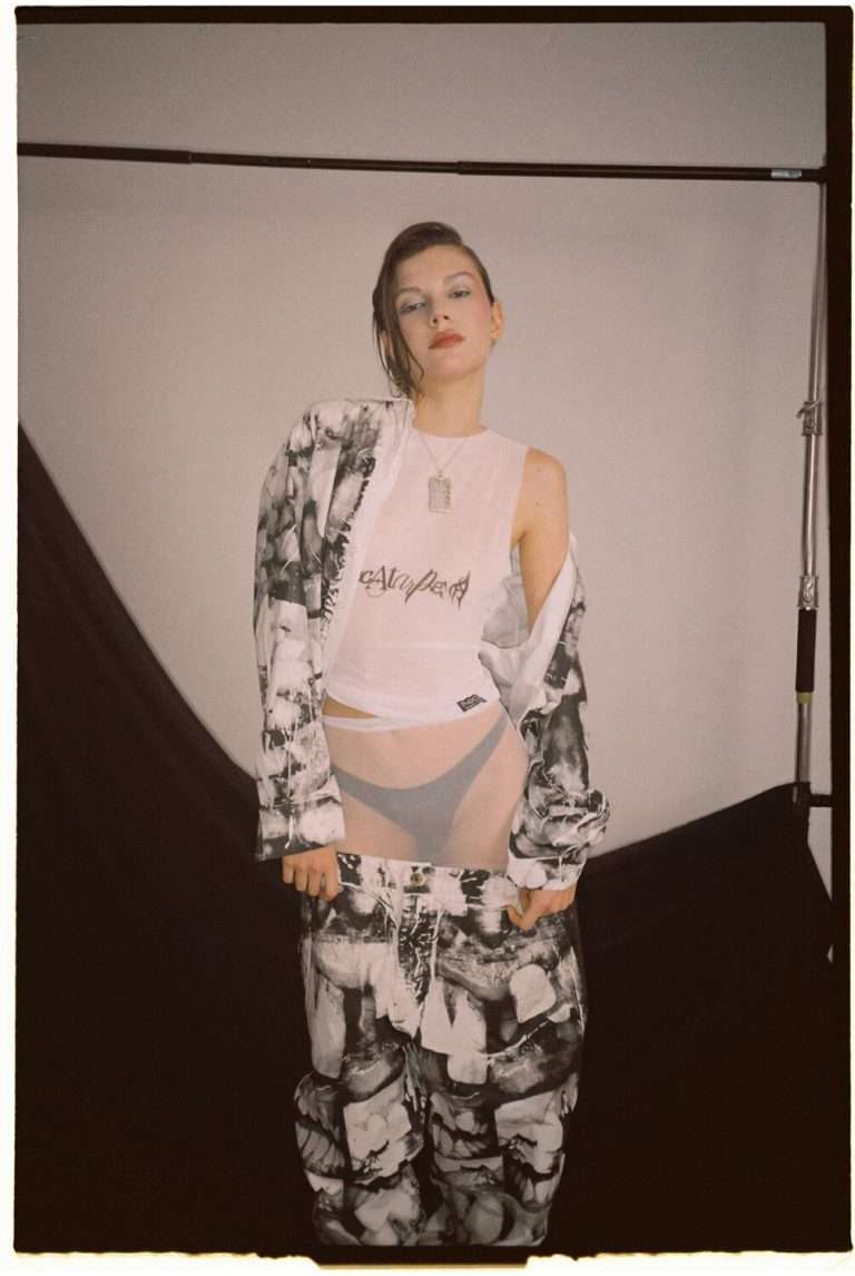 Una persona posa con confianza en un estudio, vestida con una chaqueta y pantalones estampados en blanco y negro sobre un top de malla blanca transparente y ropa interior. El fondo es sencillo, con tela negra drapeada.